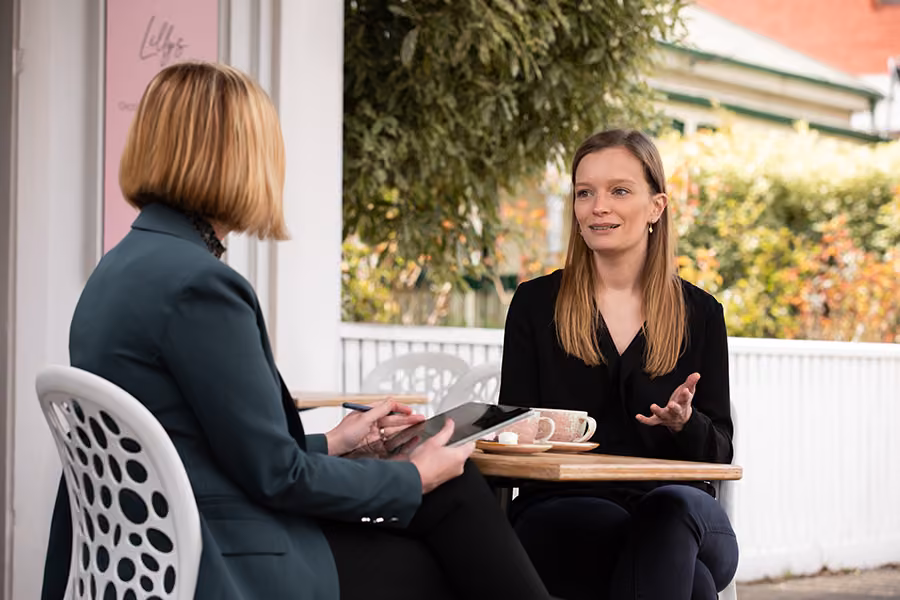 Two women facing each other, sitting outside a cafe and talking with cups of coffee in front of them on the table.