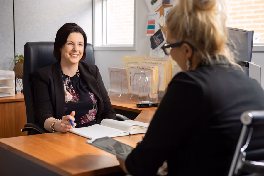Ange is sitting at her brown desk smiling at a woman sitting across from her.