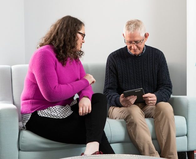 Kate is sitting on a couch in a bright pink jumper showing a man the form to complete on a tablet.