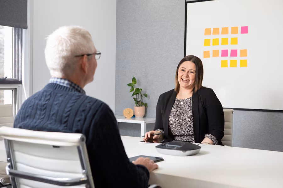 Taryn is sitting in front of a whiteboard of coloured sticky notes in the Inspire HQ boardroom talking to a man opposite her.
