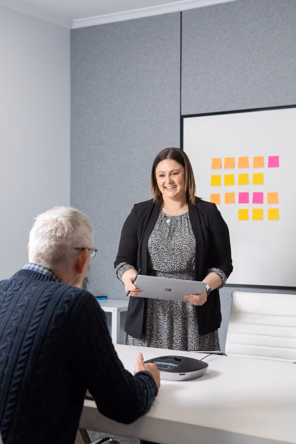 Taryn is standing, holding a computer tablet in front of a whiteboard with coloured sticky notes smiling at a man sitting across from her.