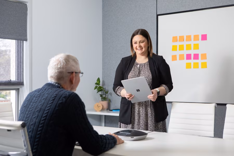 Taryn is standing in front of a whiteboard covered in colourful sticky notes while she talks and smiles at a man sitting across from her.