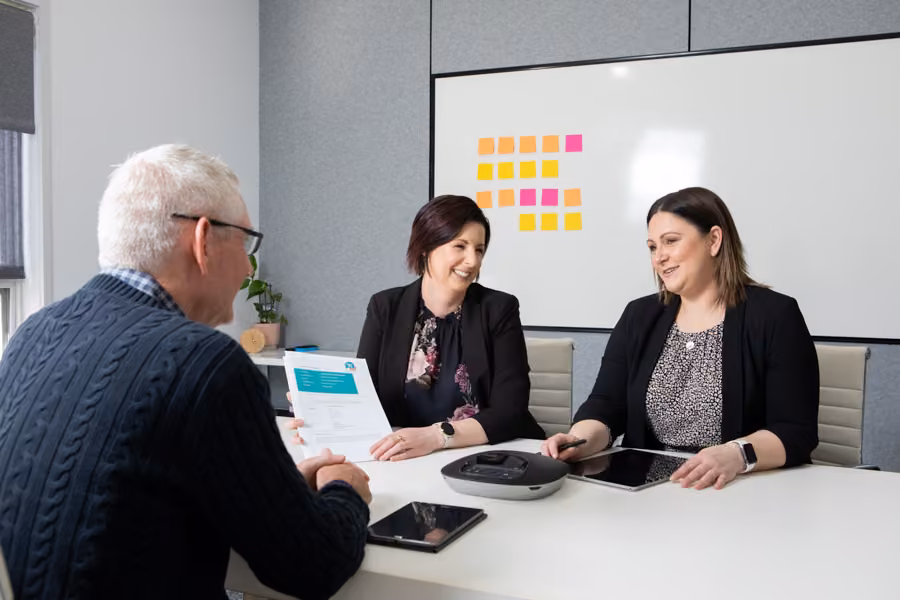 Ange is looking at Taryn while she talks to the man sitting across from them with a whiteboard of coloured sticky notes in the background.