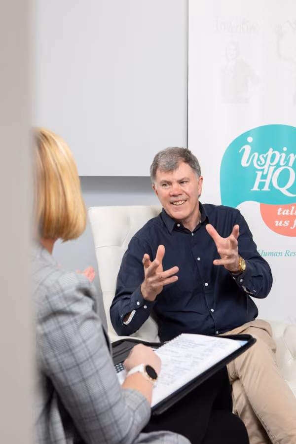 A man is sitting and gesturing with his hands as he talks to a woman sitting across from him in front of an Inspire HQ banner.