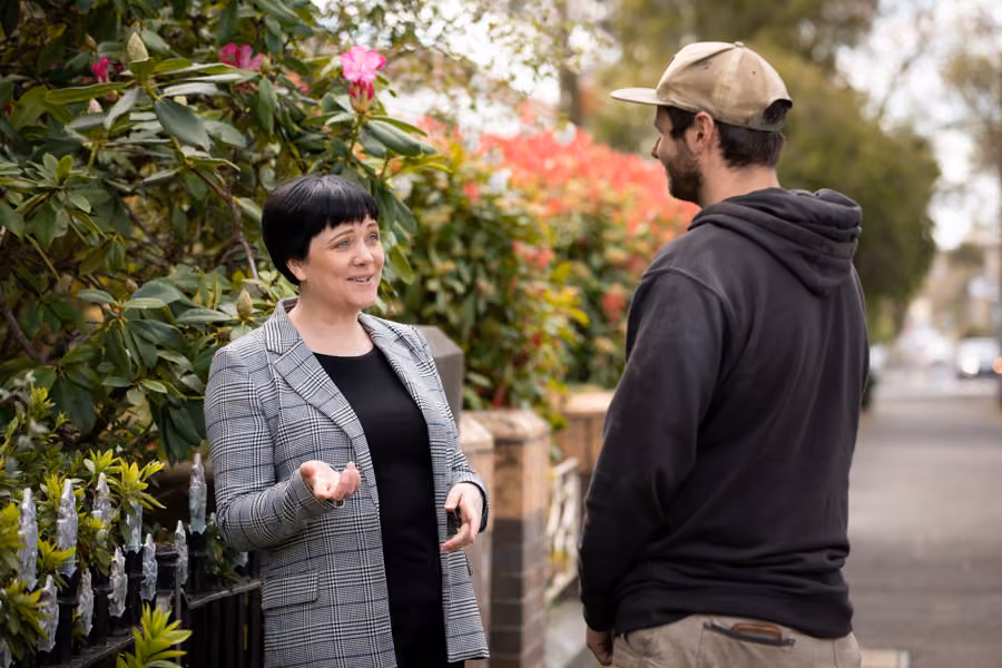 Kara is outside standing in front of a garden talking to a man.