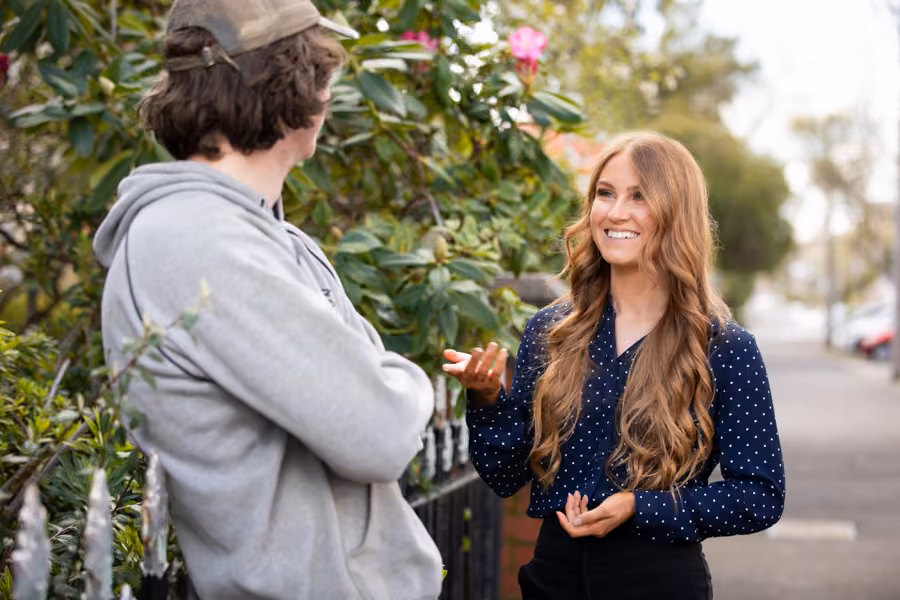 A woman with long red hair is outside talking to a man leaning against a fence.