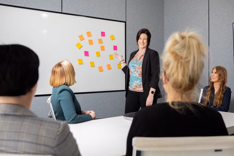 Ange is standing in front of a whiteboard with coloured sticky notes in the boardroom and presenting to Inspire HQ team members.
