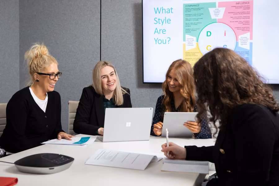 Group of women sitting around a boardroom table working with their laptops and DiSC on the tv in the background.,