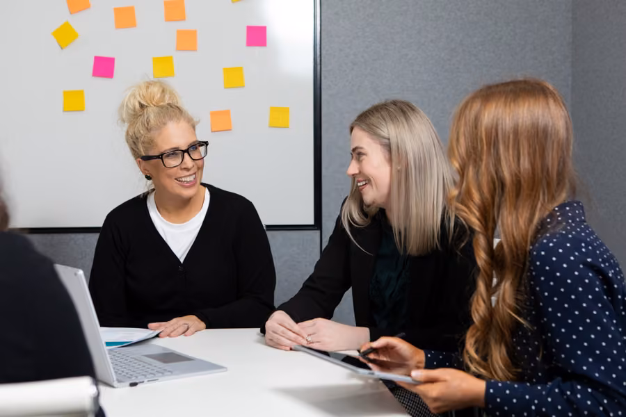 Three women are sitting around a table talking and smiling at each other with a laptop sitting on the table in front of them.