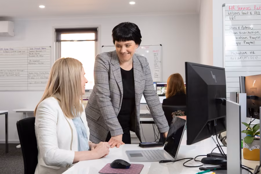 Kara is standing over a woman sitting in front of a computer smiling at each other in an office environment.